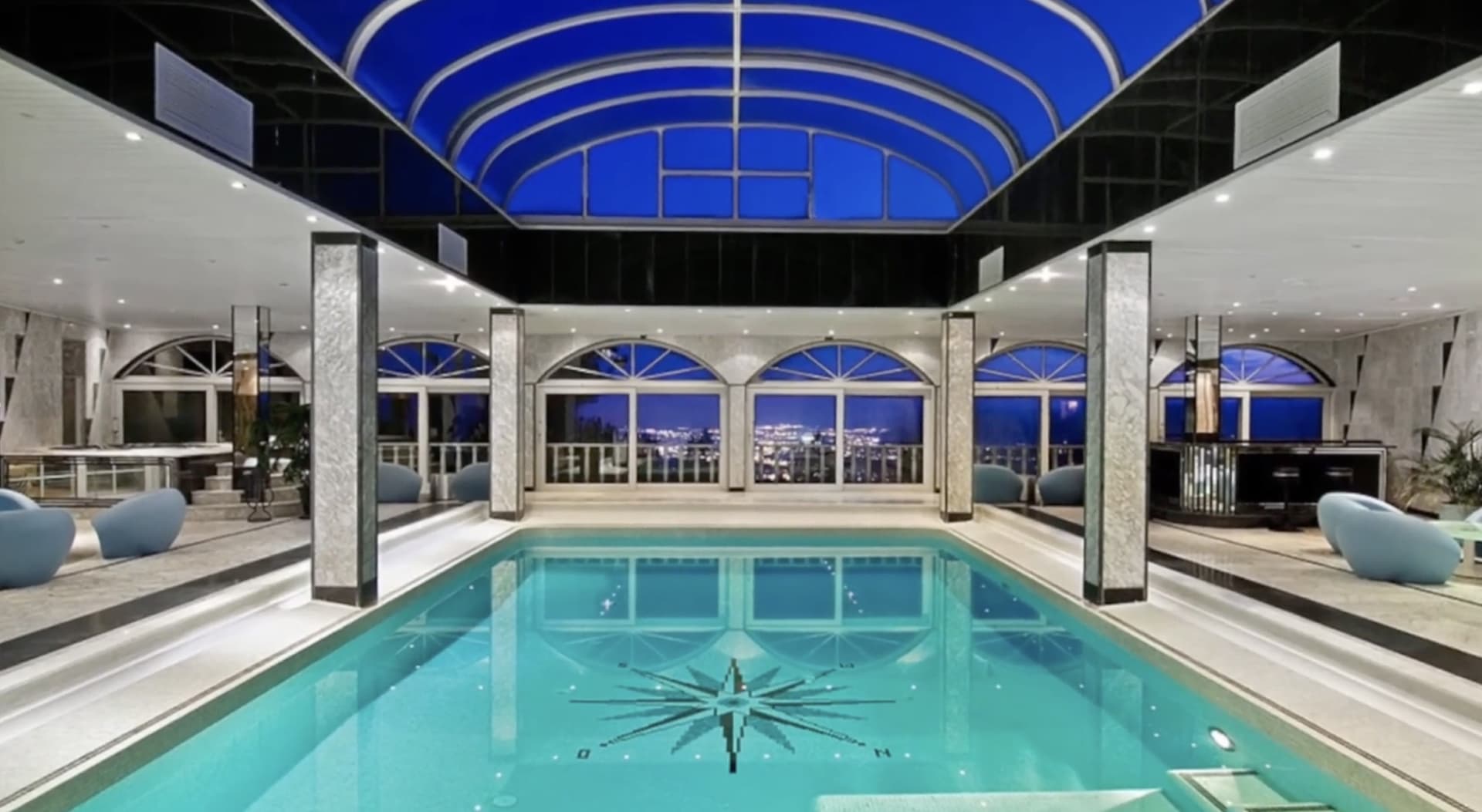 Indoor swimming pool under a barrel vaulted glass roof at dusk, arched windows looking over the lights of Ibiza