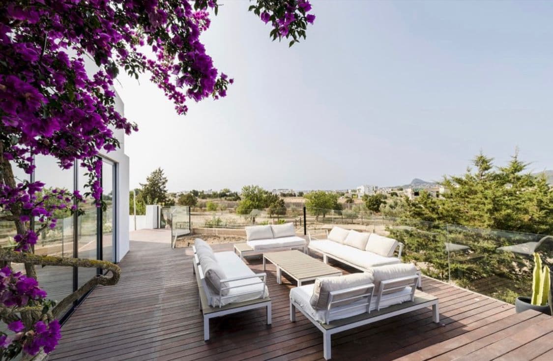 Outdoor lounge terrace with white sofas and wooden decking, framed by purple bougainvillea blooms and countryside views.