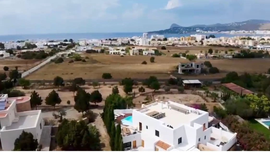 Aerial view of the villa's white rooftop terraces and pool near Sant Josep, with the Ibiza coastline and hills in the distance.