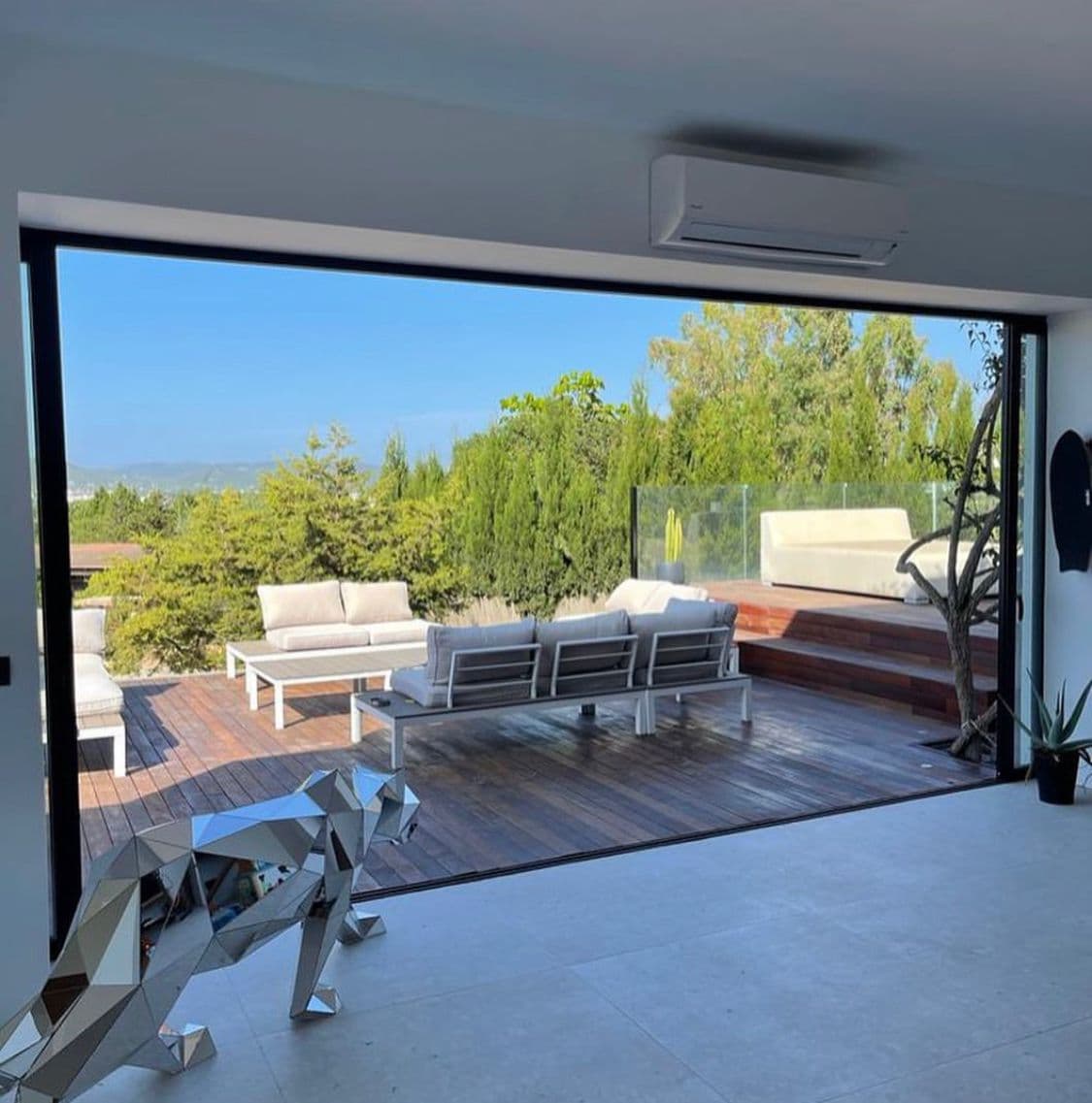 Living area with full width glass doors opening onto a wooden deck and lounge seating, surrounded by greenery.
