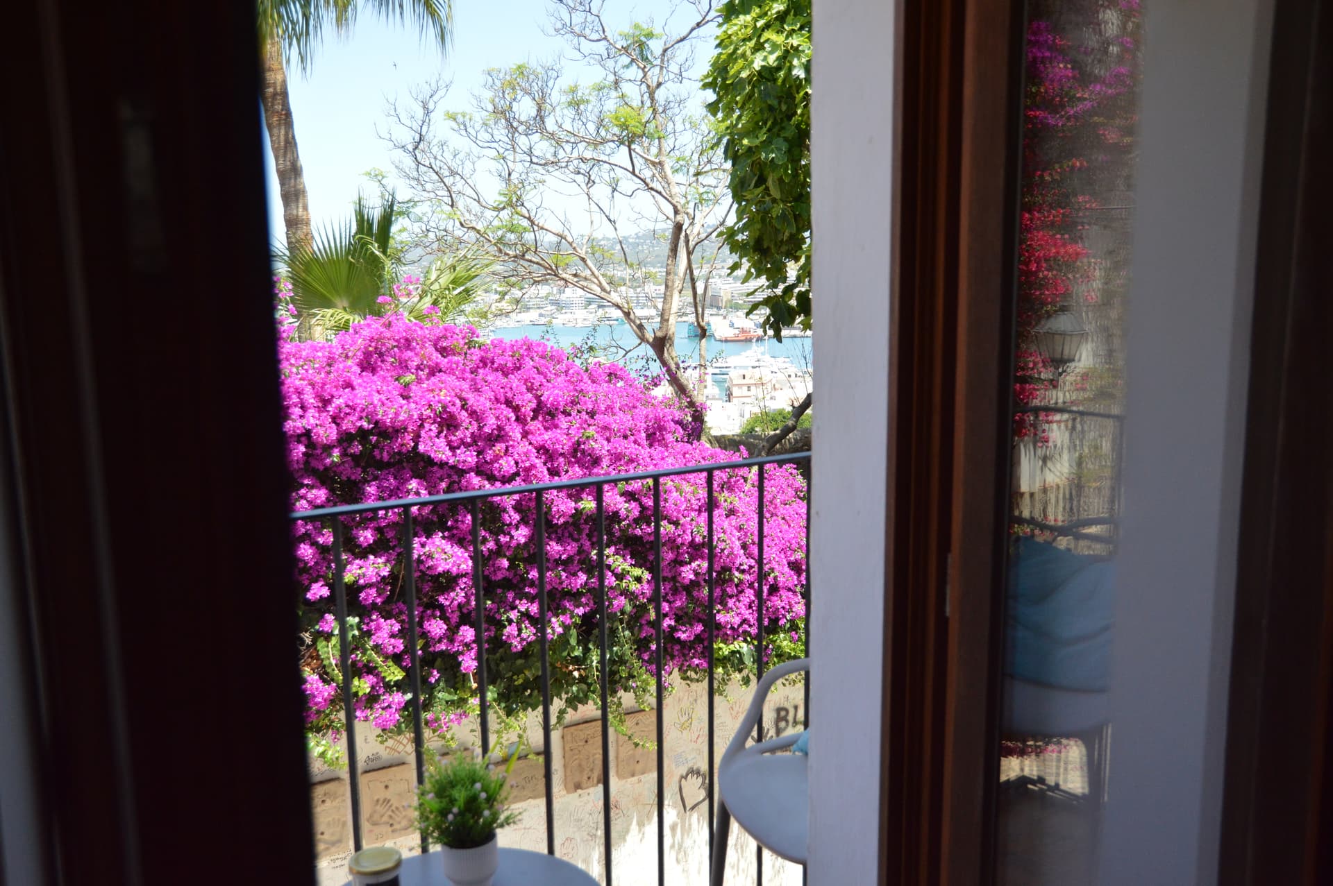 Small private balcony at Casa Mor with a table, chair and potted plant, framed by pink bougainvillea and a glimpse of the harbour beyond.