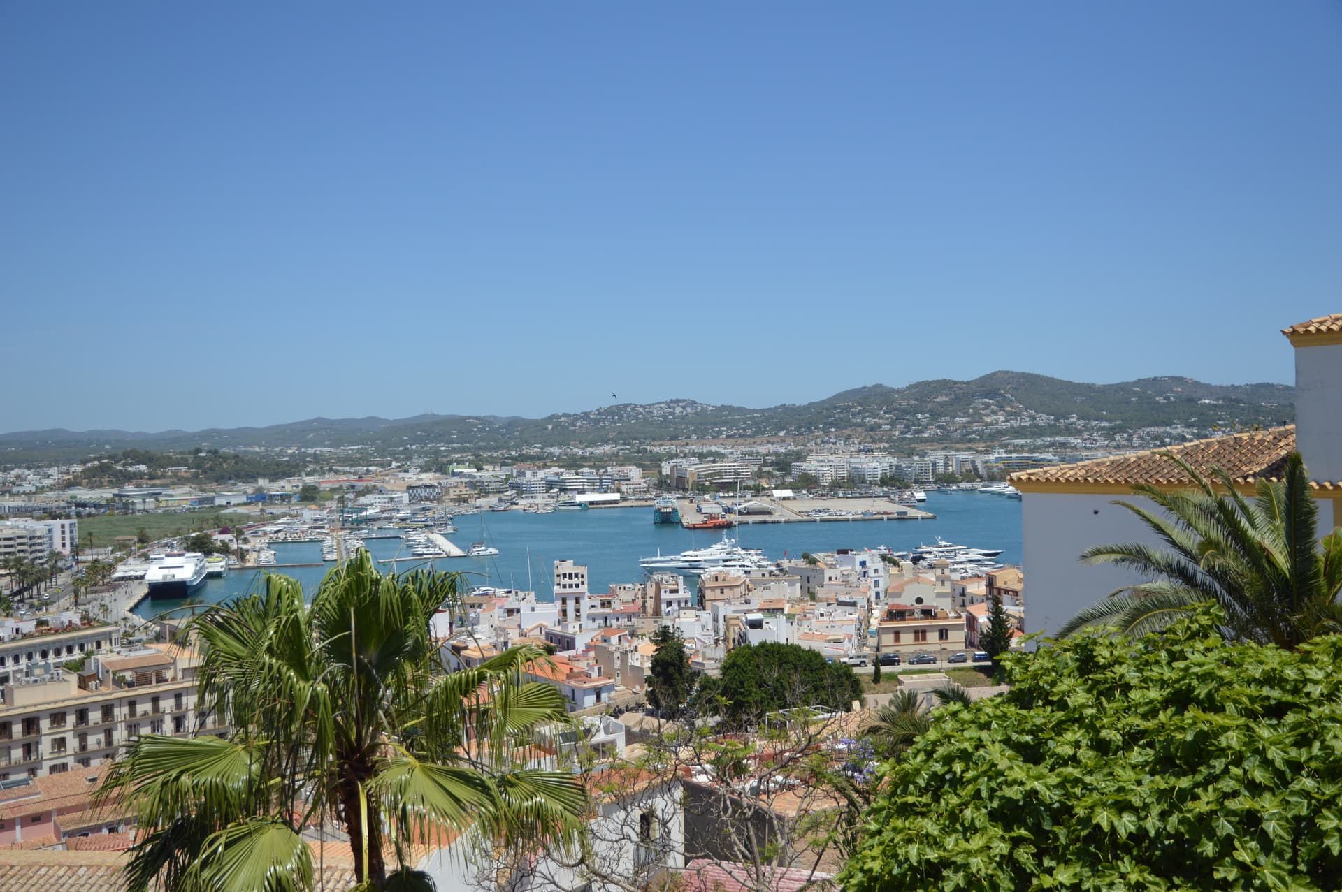 Elevated view over Eivissa harbour and rooftops from Casa Mor, showing moored boats, palm trees and distant hills under a clear sky.