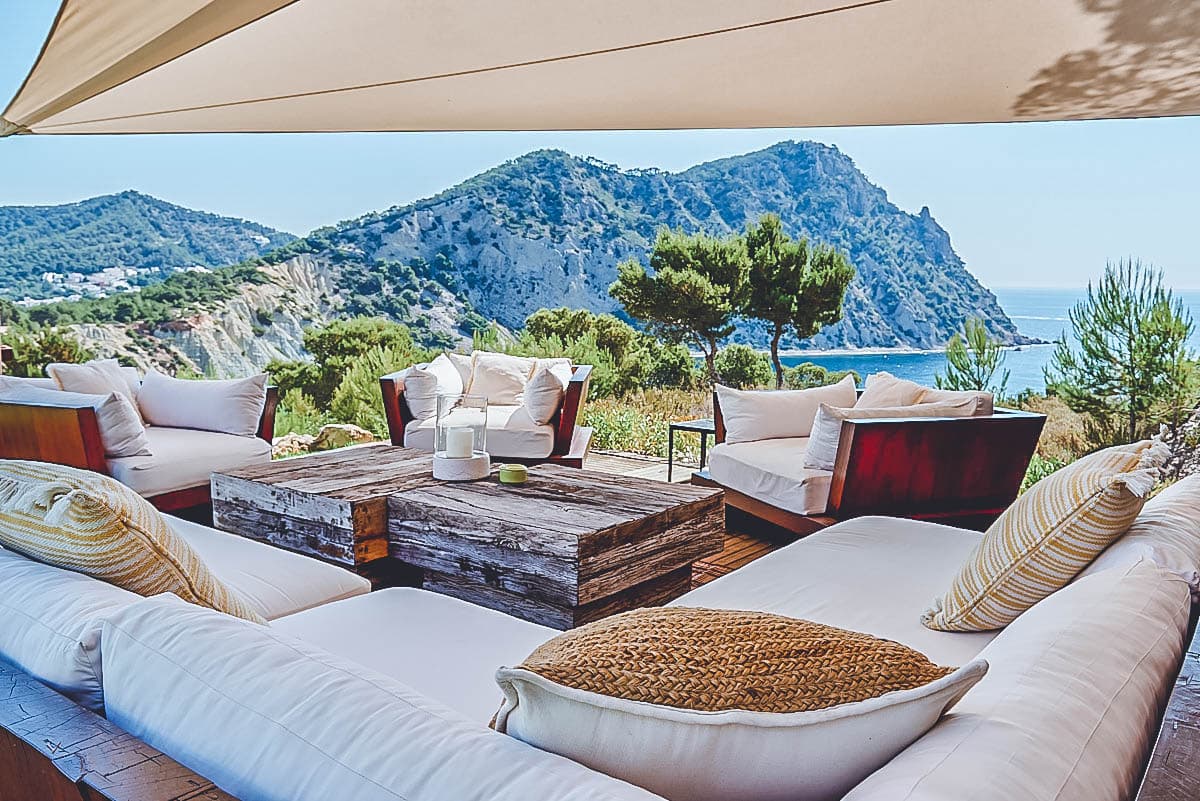 Covered outdoor lounge with deep white sofas and rustic timber table, looking out to the cliffs and sea, Sa Genista