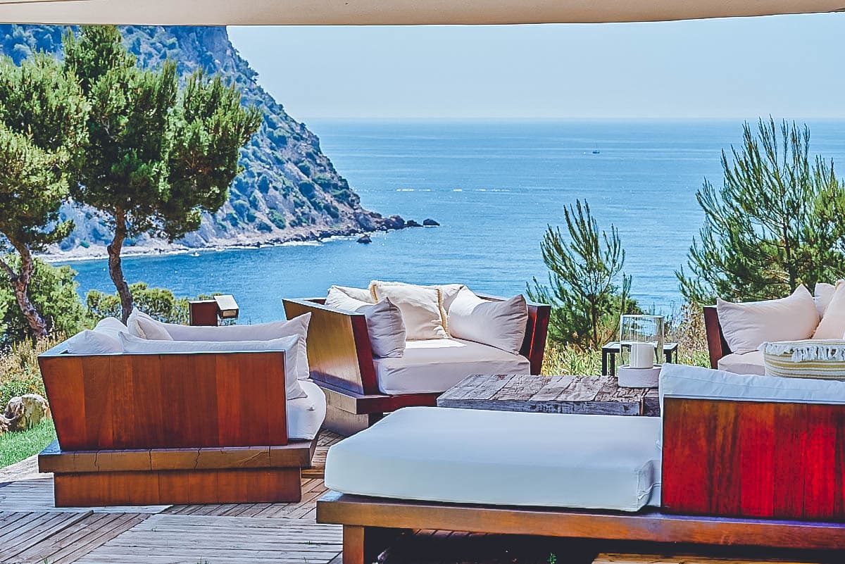 Teak terrace sofas and daybeds on a timber deck overlooking pine trees and the open sea, Sa Genista, Cala Llonga