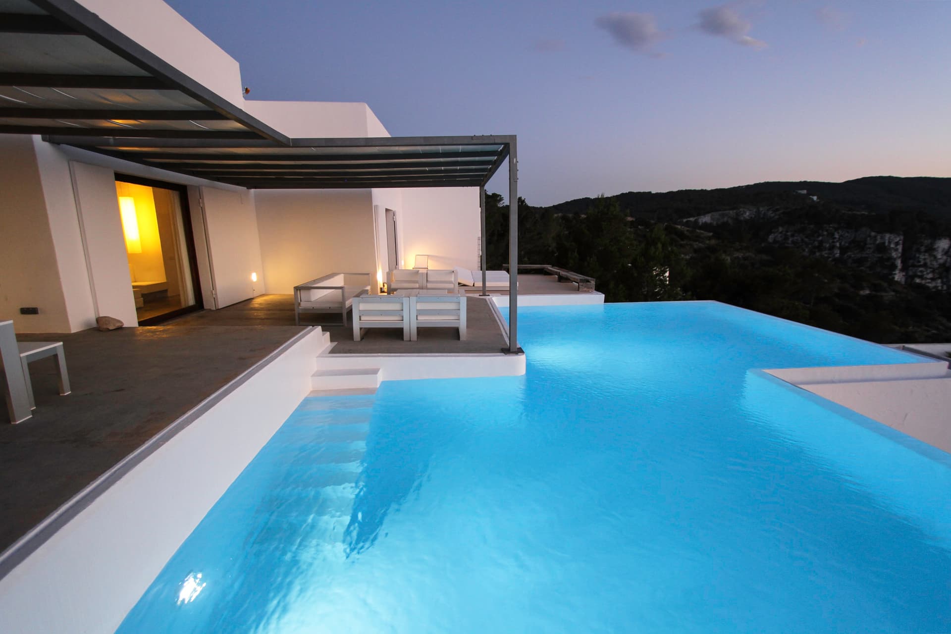 Swimming pool at dusk with the living spaces lit within at Villa Na Xamena, Sant Joan de Labritja, Ibiza