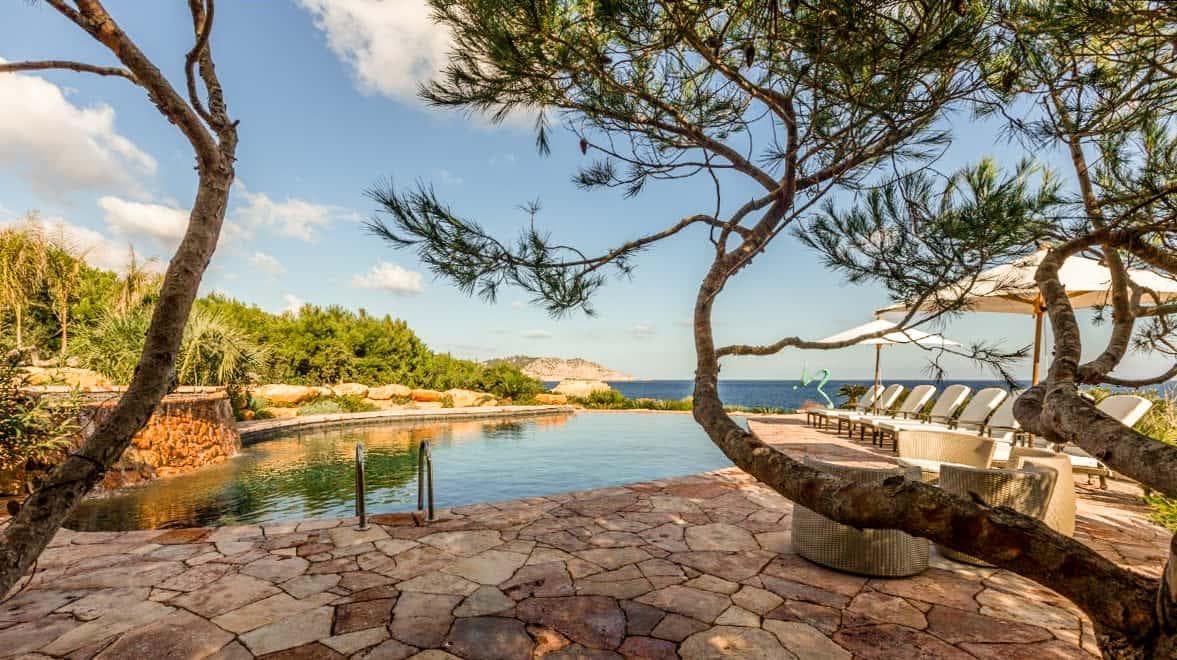 Stone terraced infinity pool at Sa Orquidea villa in Playa es Figueral, Ibiza, shaded by pine trees with sea and coastline in the distance.